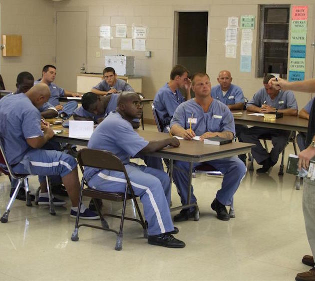 Inmates in a classroom setting, all with books and pencils, looking attentively at the instructor in the front of the room.