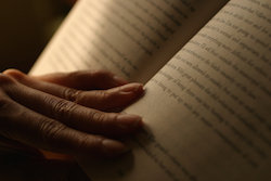 Person's hand holding a book open by the spine, text visible on the pages.