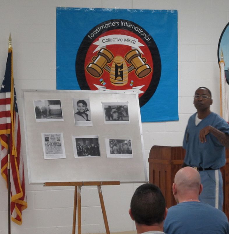 Inmate delivering a presentation, a board with pictures set up beside him, and the Toastmasters logo behind him.